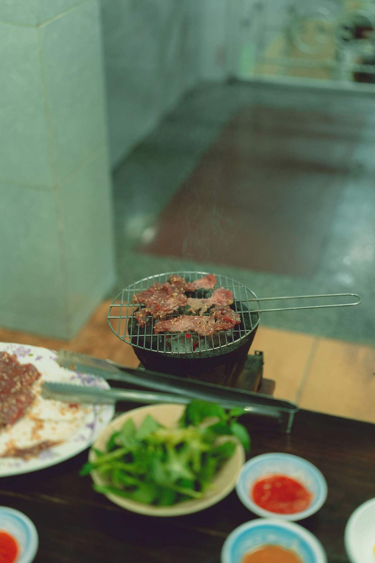 Sesame-marinated beef grilling on a wire rack over a glowing charcoal brazier on a wooden table in Đà Lạt, with a bowl of fresh herbs, a plate of raw meat and small dishes of dipping sauce