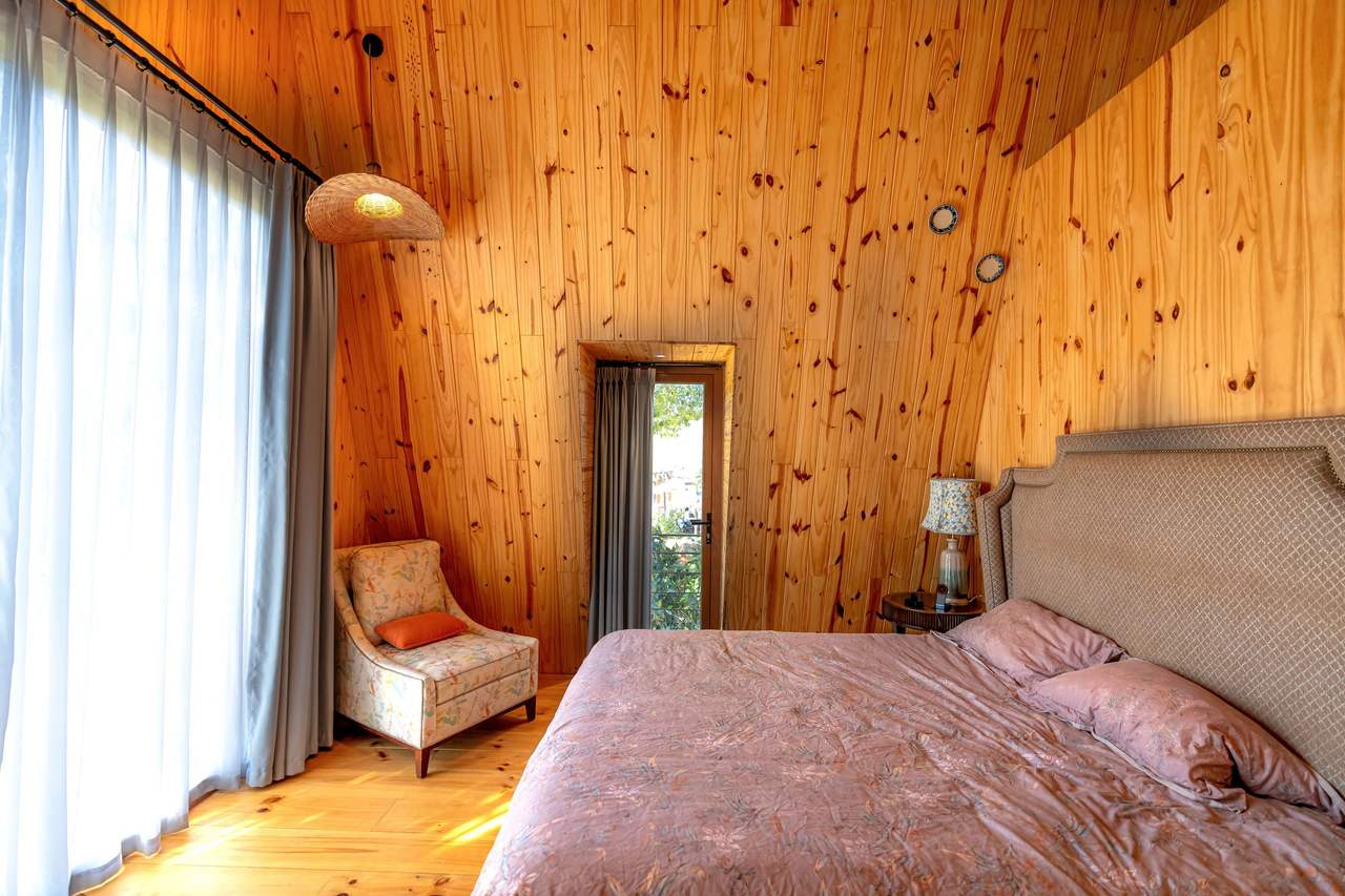 A pine-lined bedroom: a double bed with a pink cover, a small armchair, a narrow window and bright sheer curtains on the left, sun on the wooden floor
