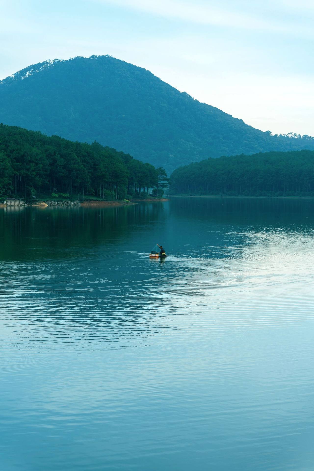 A still blue lake in Đà Lạt in the morning, one person paddling a small boat in the middle, pine hills and a mountain behind