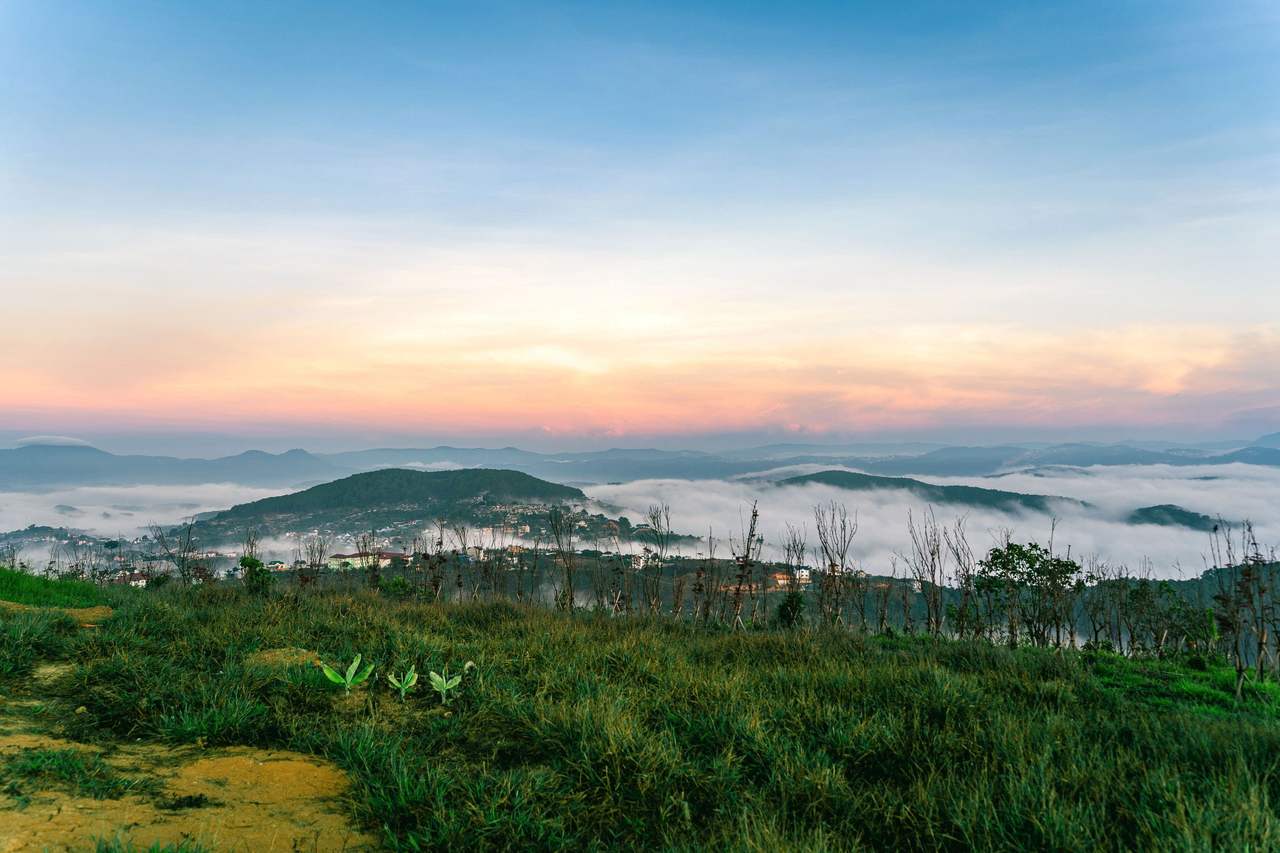A green grassy hill in Đà Lạt at daybreak above a sea of cloud filling the valley, a few hilltops rising out of it in the distance