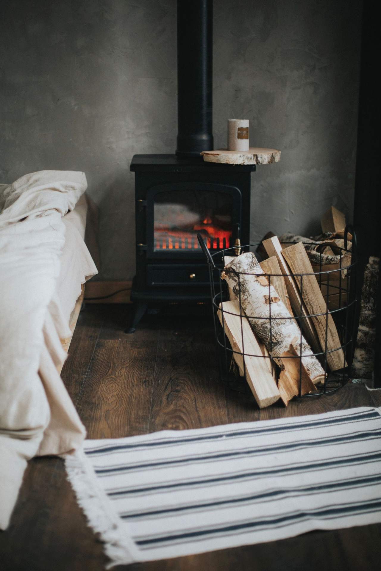 A black cast-iron stove with a small fire behind its glass door, a basket of birch logs beside it, the edge of a white bed and a striped rug in the foreground