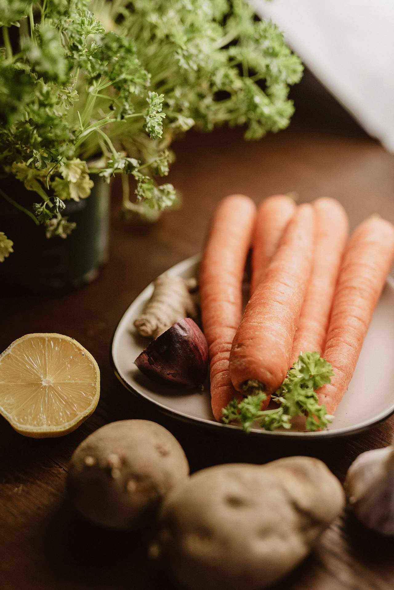 Carrots, potatoes, a lemon and fresh greens laid out on a dark wooden kitchen table in slanting window light