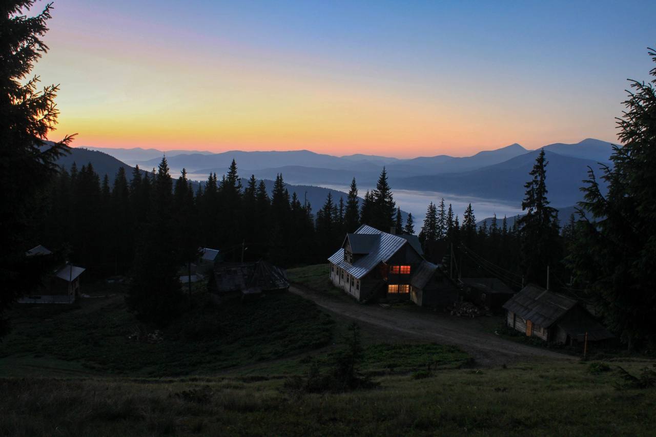A two-storey wooden house at blue hour among conifers: two windows glowing warm orange, a metal roof catching the blue light, and behind it a cloud-filled valley and mountain ridges under the last band of twilight