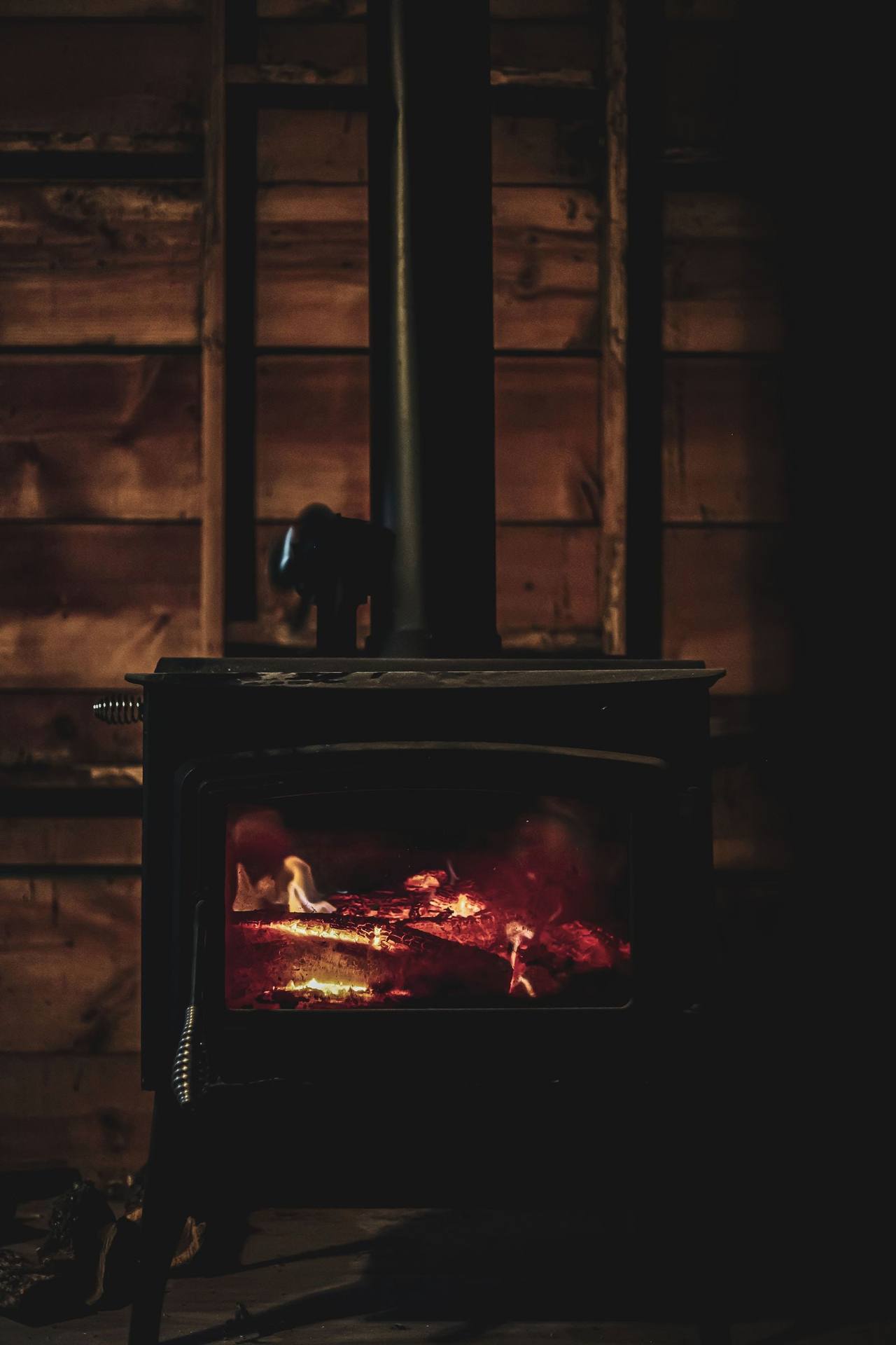 A cast-iron stove in a dark wooden room, embers and orange flames behind the glass the only light