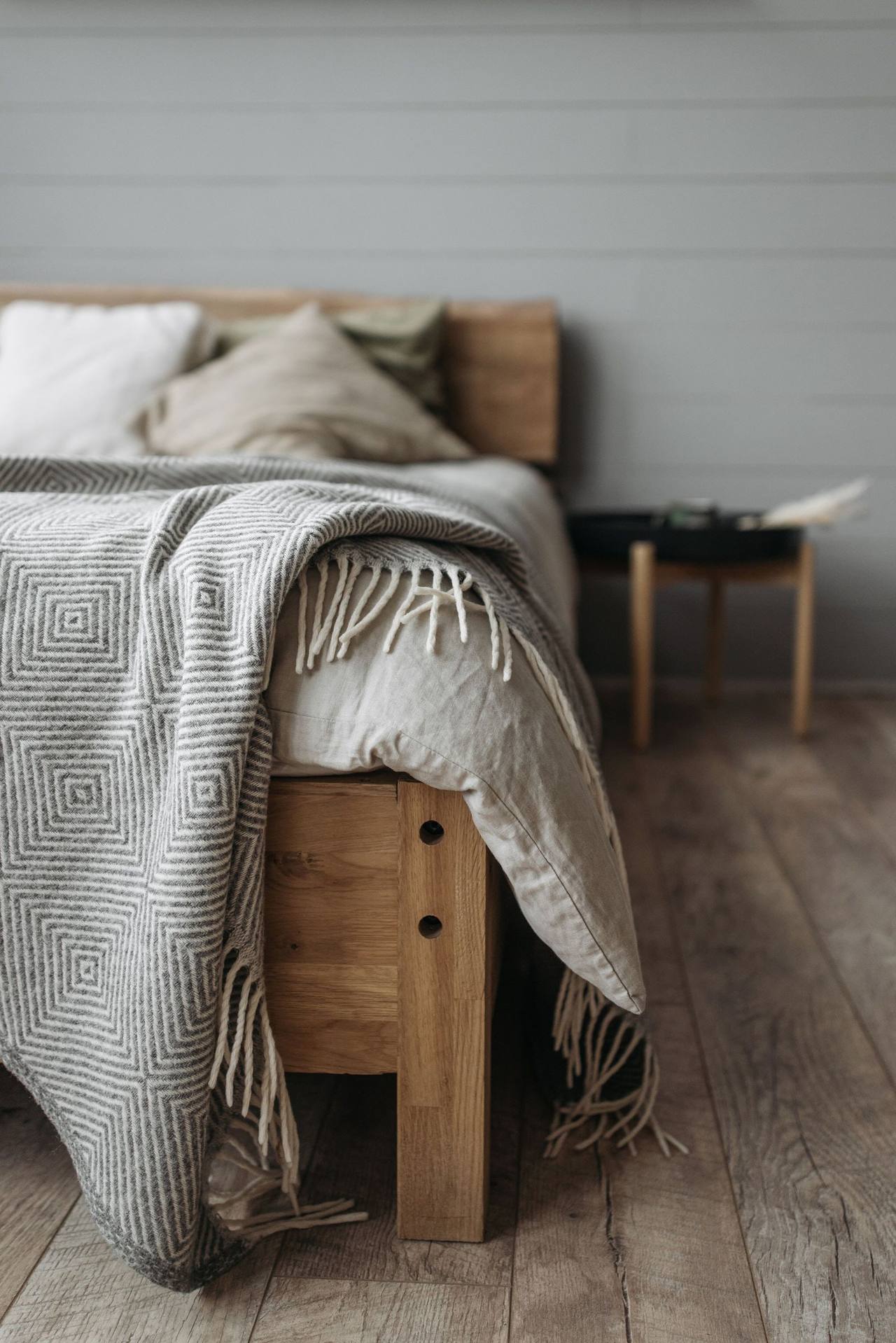 A grey fringed wool blanket draped over the edge of a wooden bed, pillows and a duvet above it, a wooden floor and a small stool beside the bed