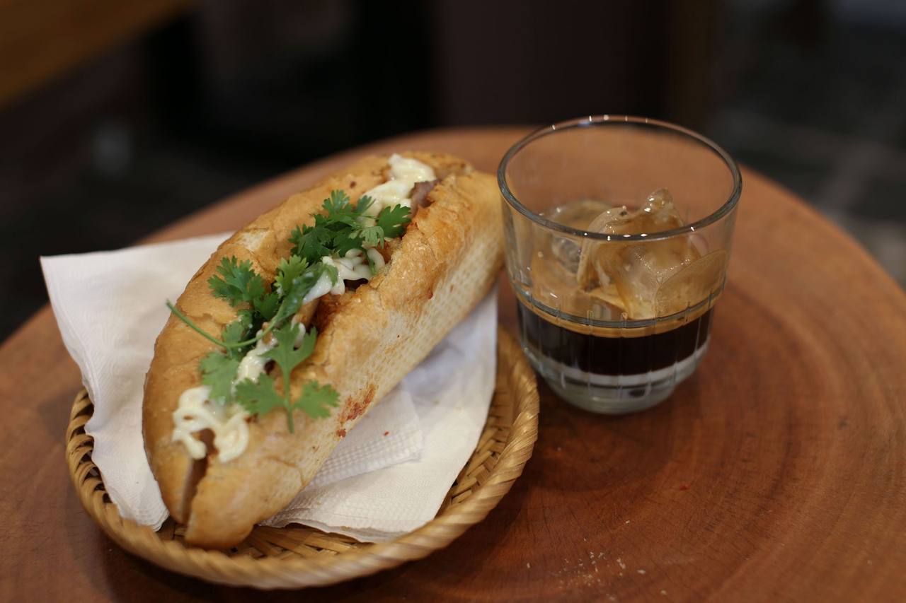 A bánh mì with coriander in a paper-lined bamboo basket next to a glass of iced coffee with milk, on a round wooden table