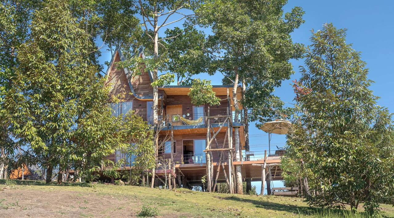 A two-storey wooden house with a steep roof and wraparound porches among tall trees on a grassy slope, under a blue sky
