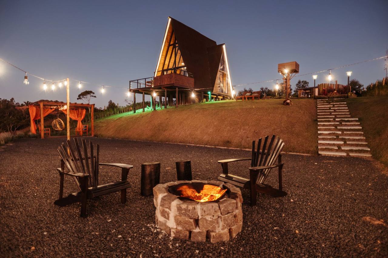 The yard at dusk: a round stone fire pit burning, four wooden chairs around it, a string of bulb lights, stone steps leading up to the wooden house on the slope