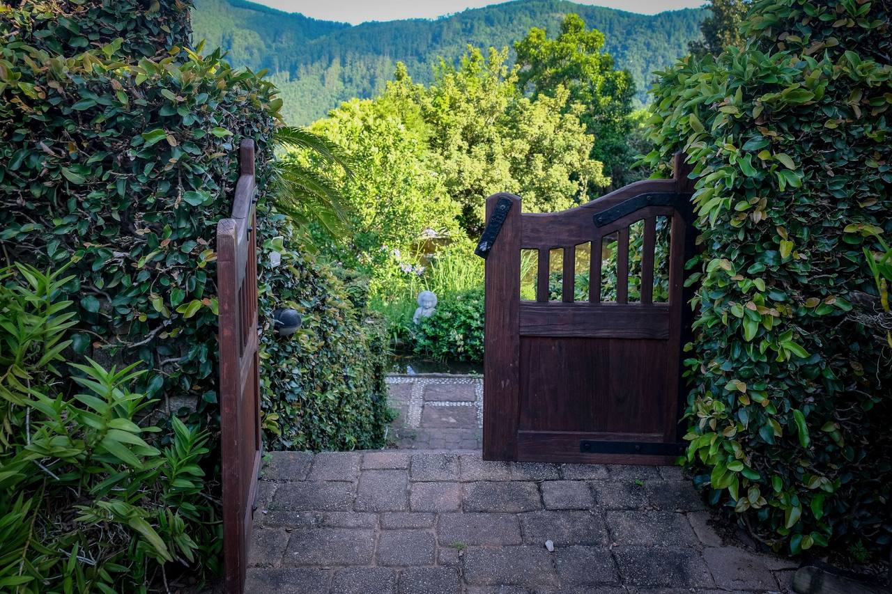 A wooden gate standing half open between two walls of climbing plants, a paved path leading into the garden, green hills behind