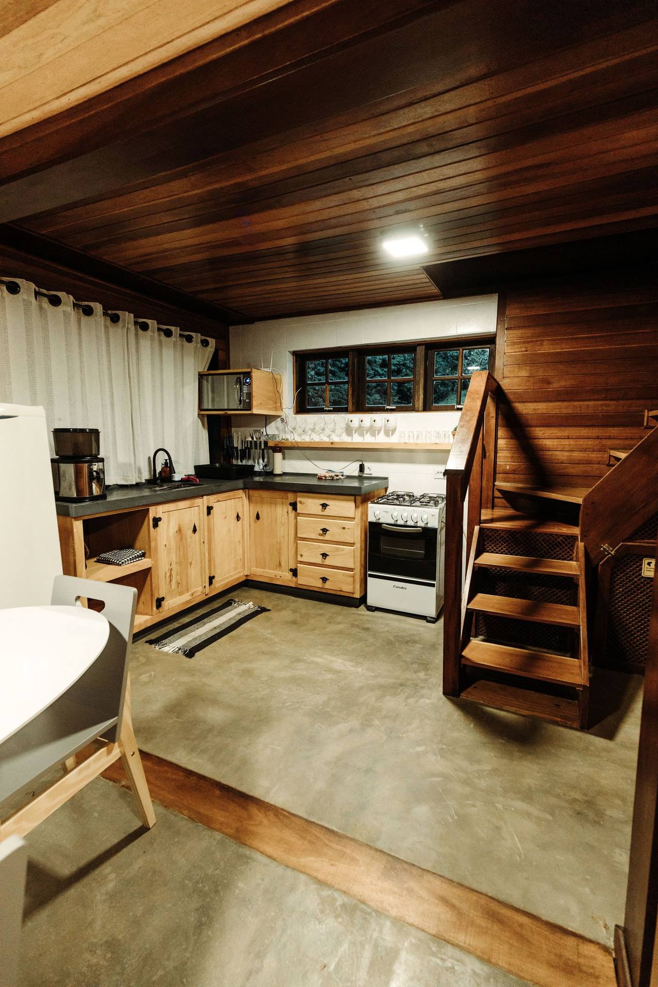 The shared kitchen under a dark timber ceiling: an L of pale wooden cabinets, a grey stone counter, a coffee maker and knife rack, a window onto trees, a wooden stair on the right