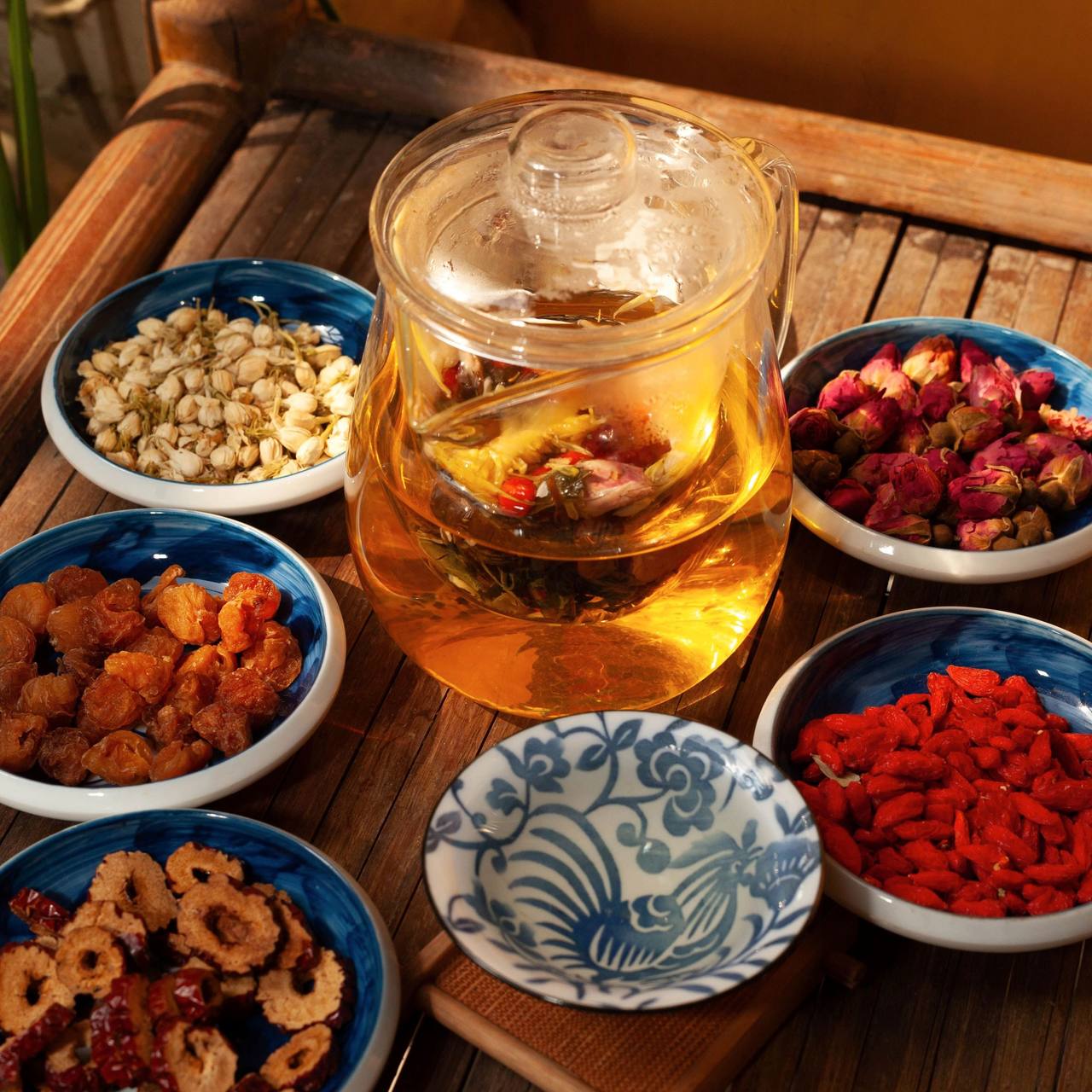 A glass teapot of golden herbal tea on a bamboo tray, surrounded by blue china bowls of dried rosebuds, red dates, goji berries and chrysanthemum