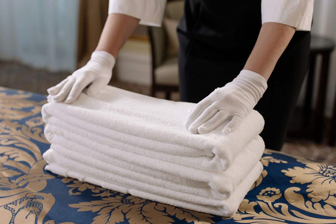 Two white-gloved hands smoothing a stack of freshly folded white towels on a patterned bedspread