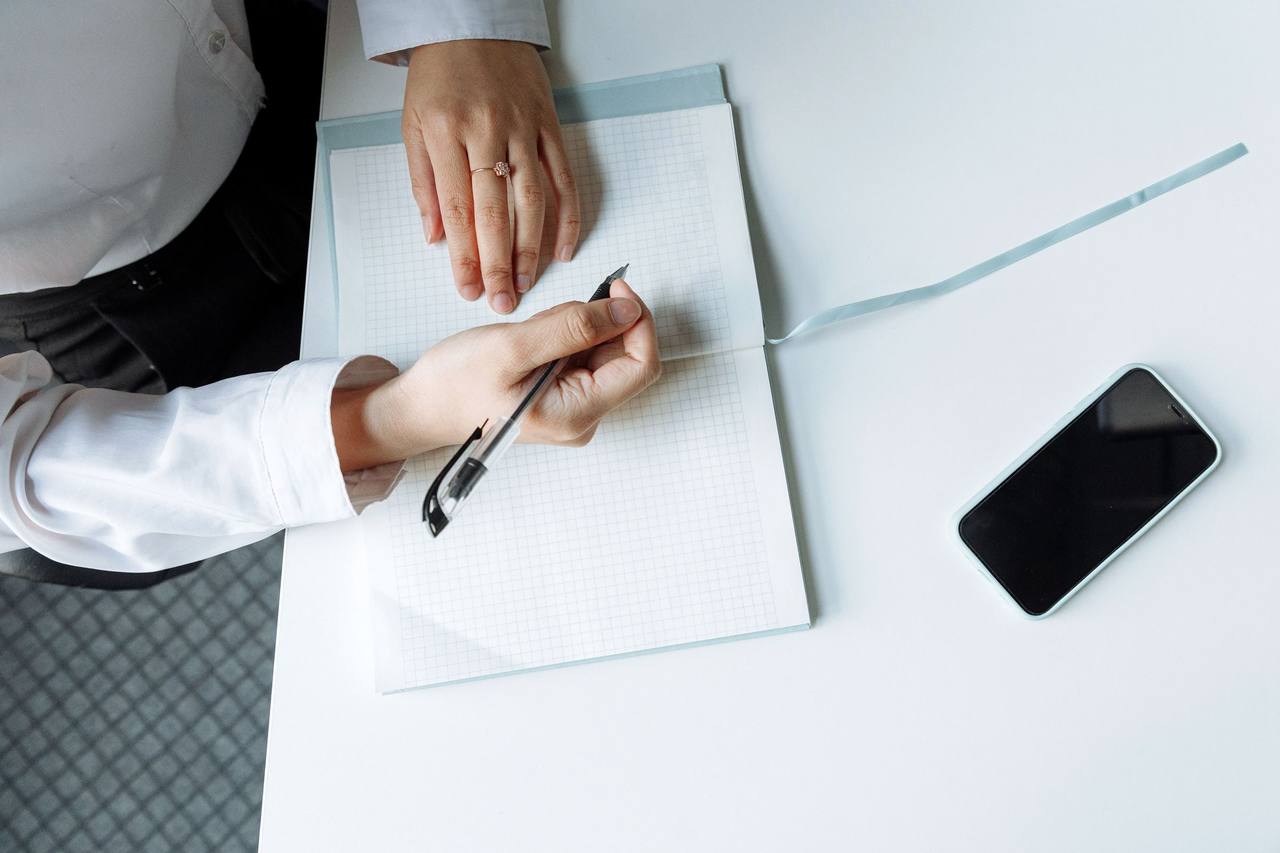 From above: two hands with a pen on a grid notebook on a white desk, a phone with a dark screen lying beside it