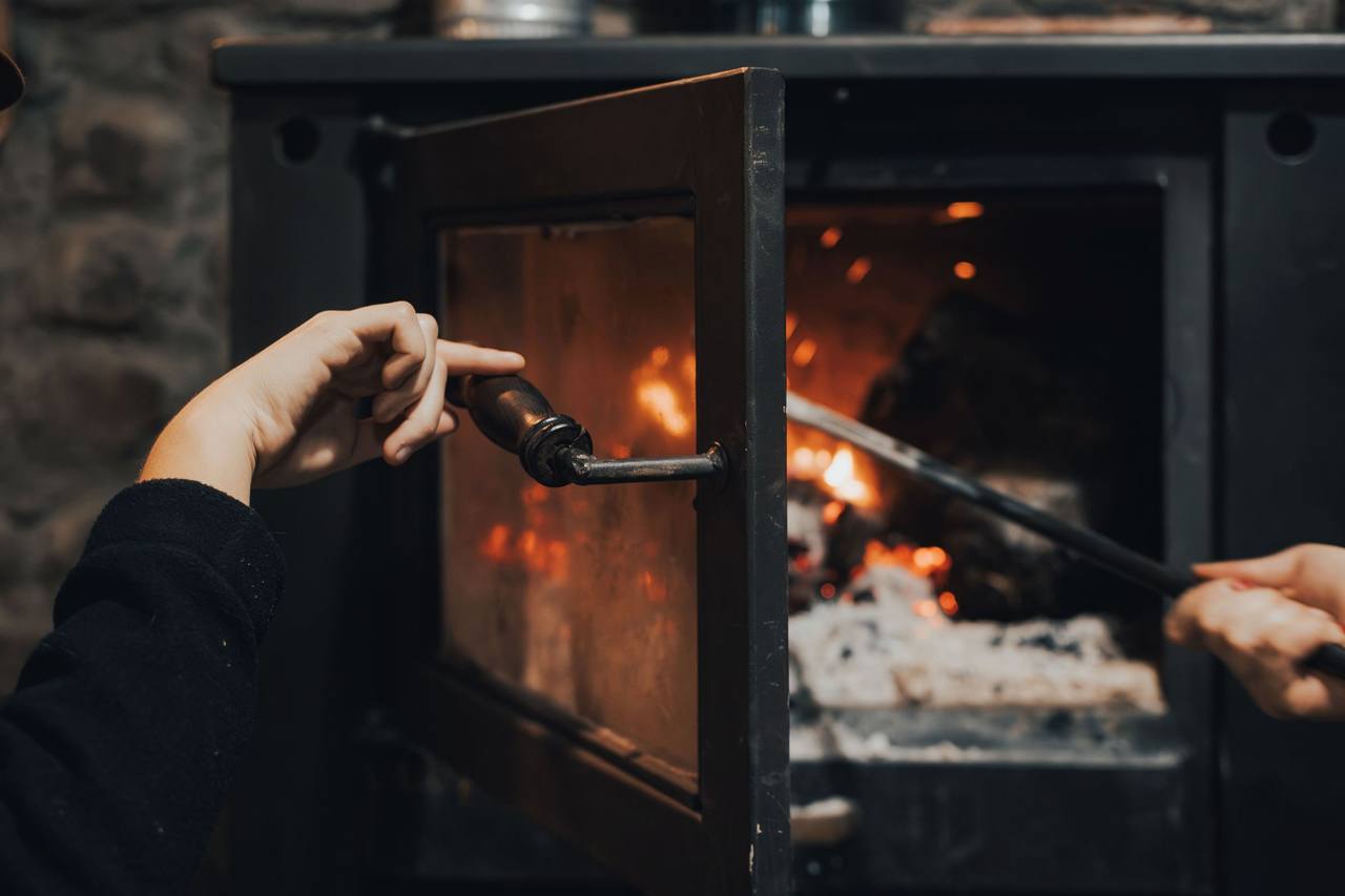 A hand opening the glass door of a cast-iron stove while the other hand adjusts the glowing logs inside with a poker