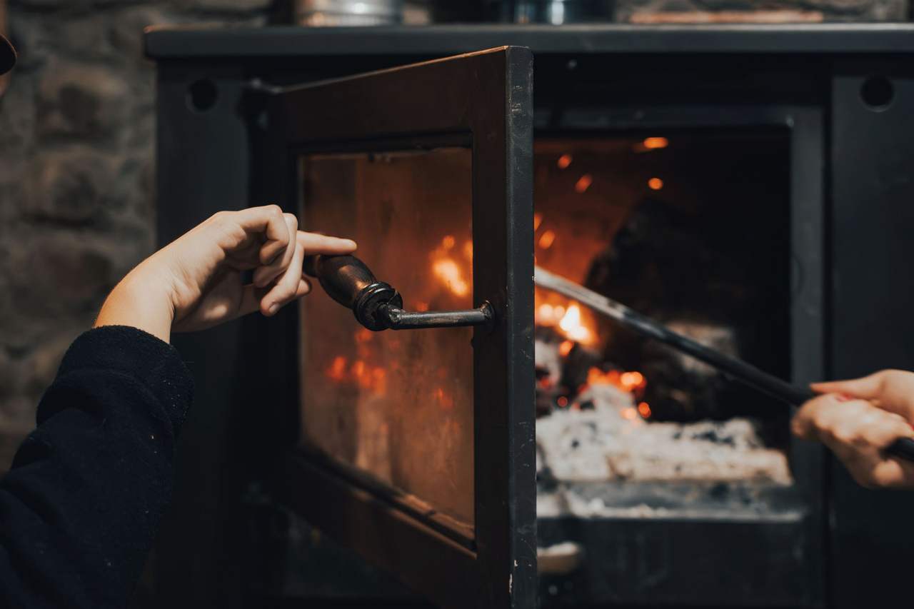 A hand opening the glass door of a cast-iron stove while the other hand adjusts the glowing logs inside with a poker