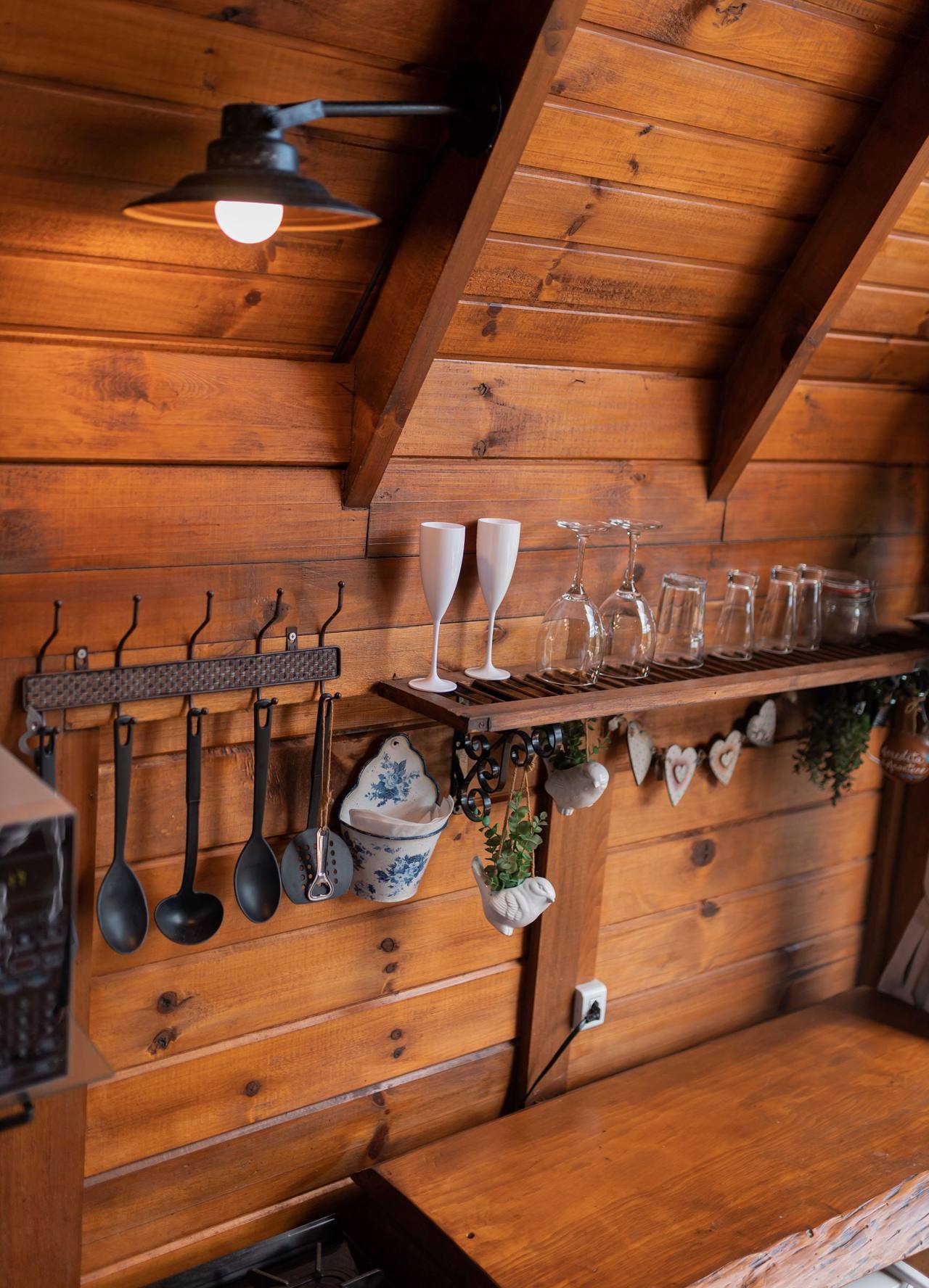 The Kitchen Room's kitchen corner: ladles hanging from an iron rail, a wooden shelf of glasses and china, a lamp under the sloping timber roof