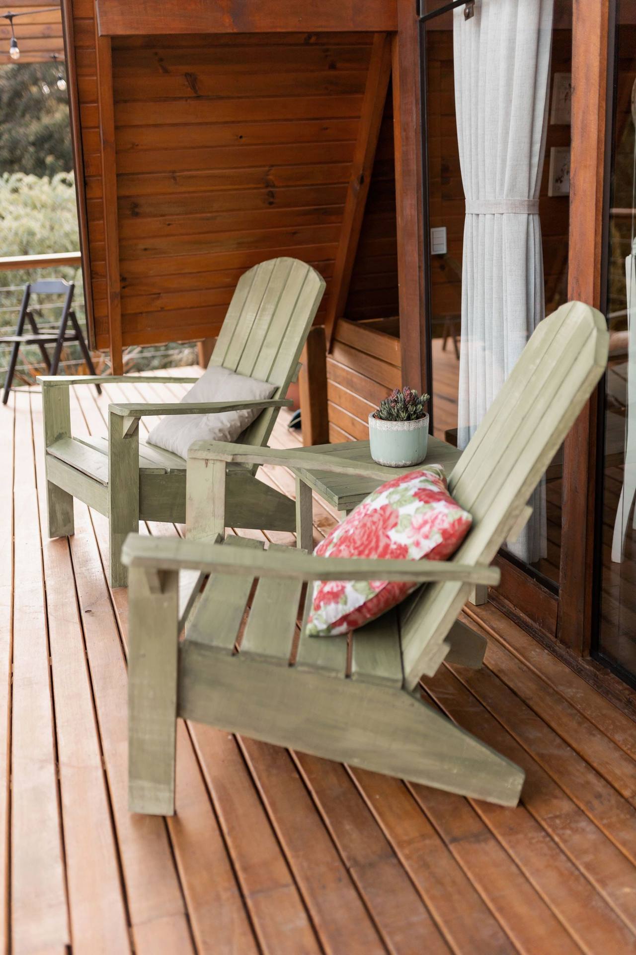The Kitchen Room terrace: two grey-green wooden lounge chairs, a red floral cushion, a small potted plant, woods beyond the railing