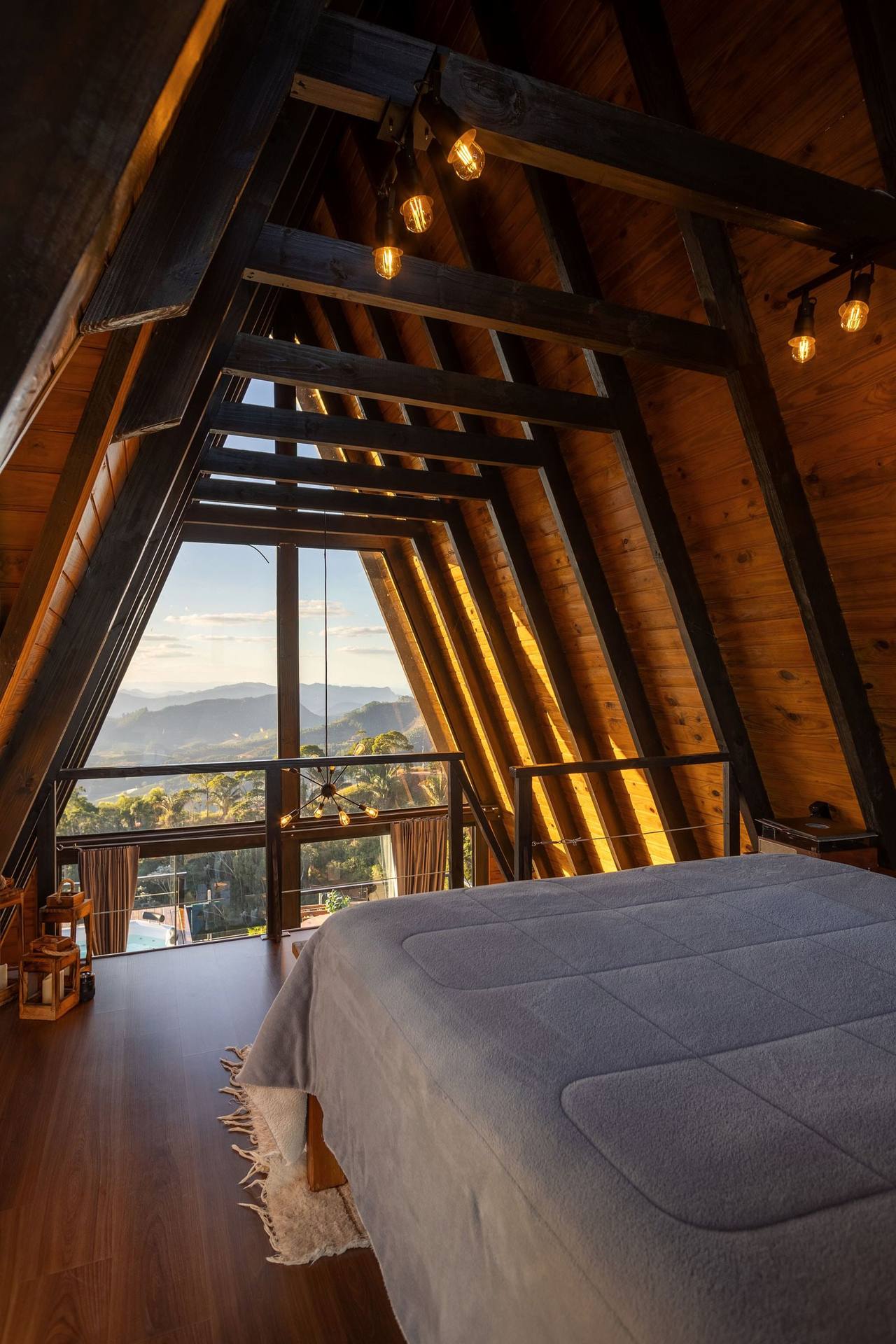 Cloud Loft, portrait view: a bed with a grey quilt under dark timber rafters, the glass gable with its railing looking out over a green valley