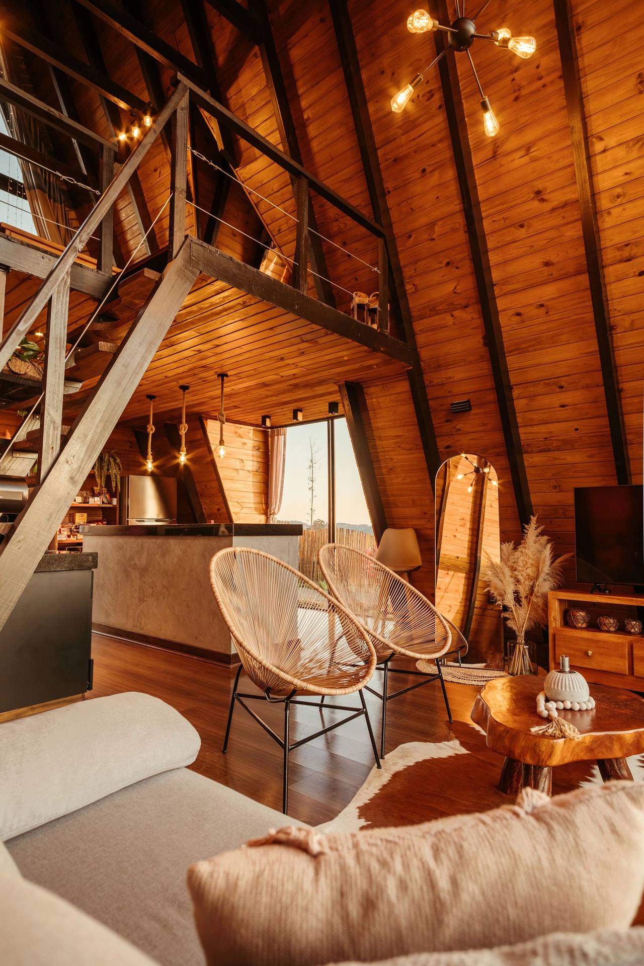 The Cloud Loft from the top: the steep timber stair to the sleeping loft on the left, two wicker chairs and a sofa below, a tall window at the far end