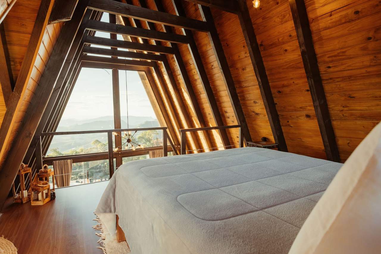 Cloud Loft: a queen bed on the loft under a wood-lined A-frame roof, the glass gable with a railing looking out over the valley and distant hills