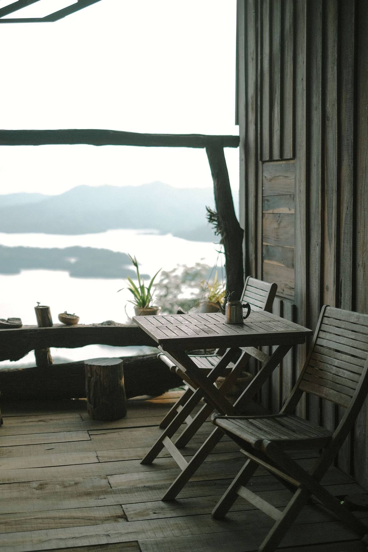 The timber porch early in the morning: a folding table and two wooden chairs against the plank wall, a small teapot on the table, a rough wooden rail looking out on hills and a misty lake