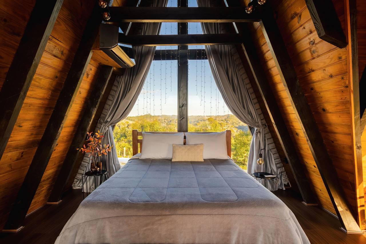 The Sun Porch bed seen straight on from its foot: grey quilt, white pillows, the glass gable between two curtains opening onto trees and hills