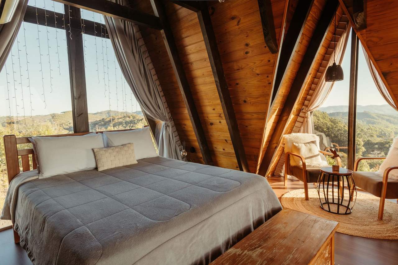 Sun Porch: a large bed with a grey quilt under a wood-lined A-frame roof, a floor-to-roof glass gable with sheer curtains on each side, an armchair by the glass, trees and hills outside in early sun