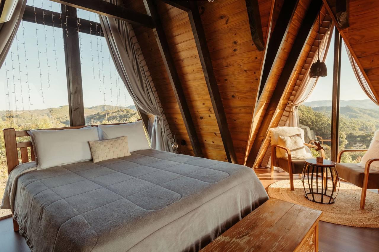 Sun Porch: a large bed with a grey quilt under a wood-lined A-frame roof, a floor-to-roof glass gable with sheer curtains on each side, an armchair by the glass, trees and hills outside in early sun