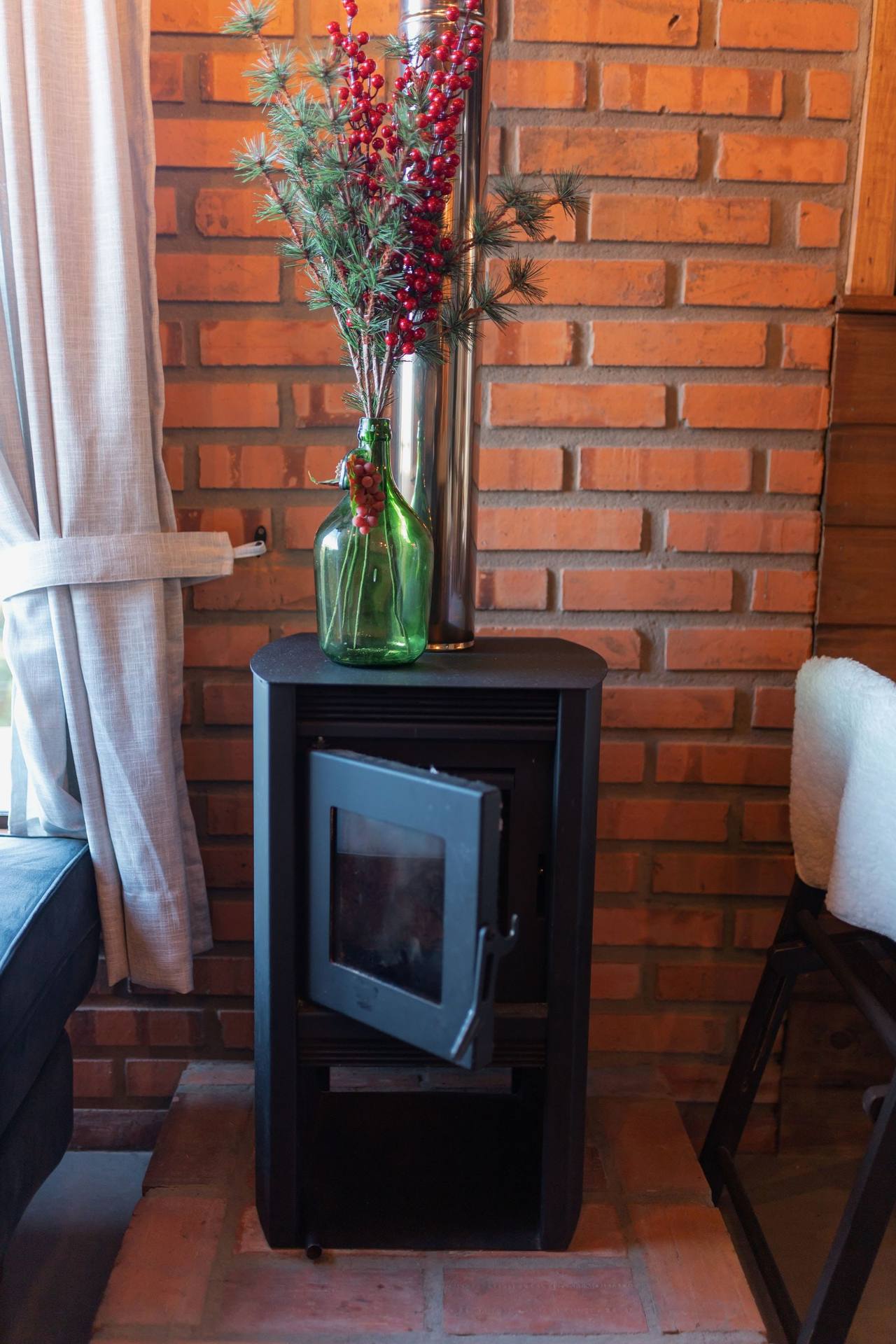 The black glass-fronted stove on a brick hearth against a bare brick wall in the Fireside room, a vase of red berries on top, white sheer curtains at the window