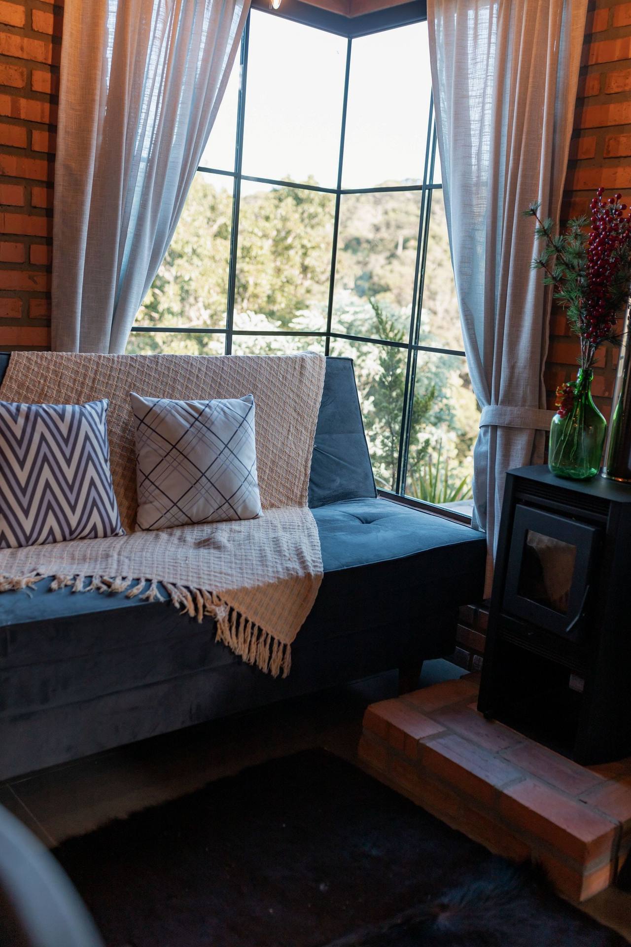 Fireside: a cushioned window seat against the corner glass looking onto the woods, a wool throw and patterned pillows, a black stove with a glass door on a brick plinth to the right