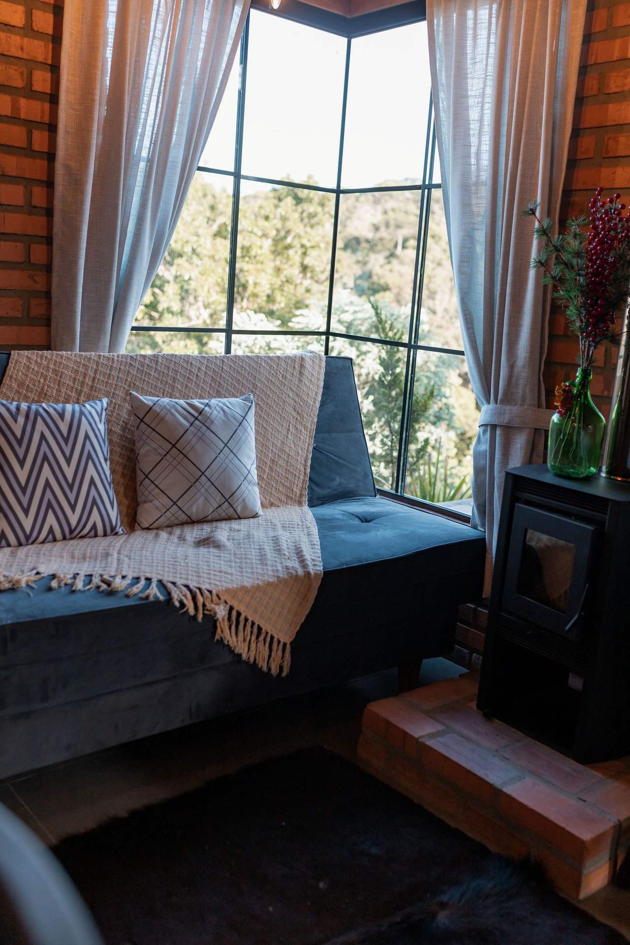 Fireside: a cushioned window seat against the corner glass looking onto the woods, a wool throw and patterned pillows, a black stove with a glass door on a brick plinth to the right