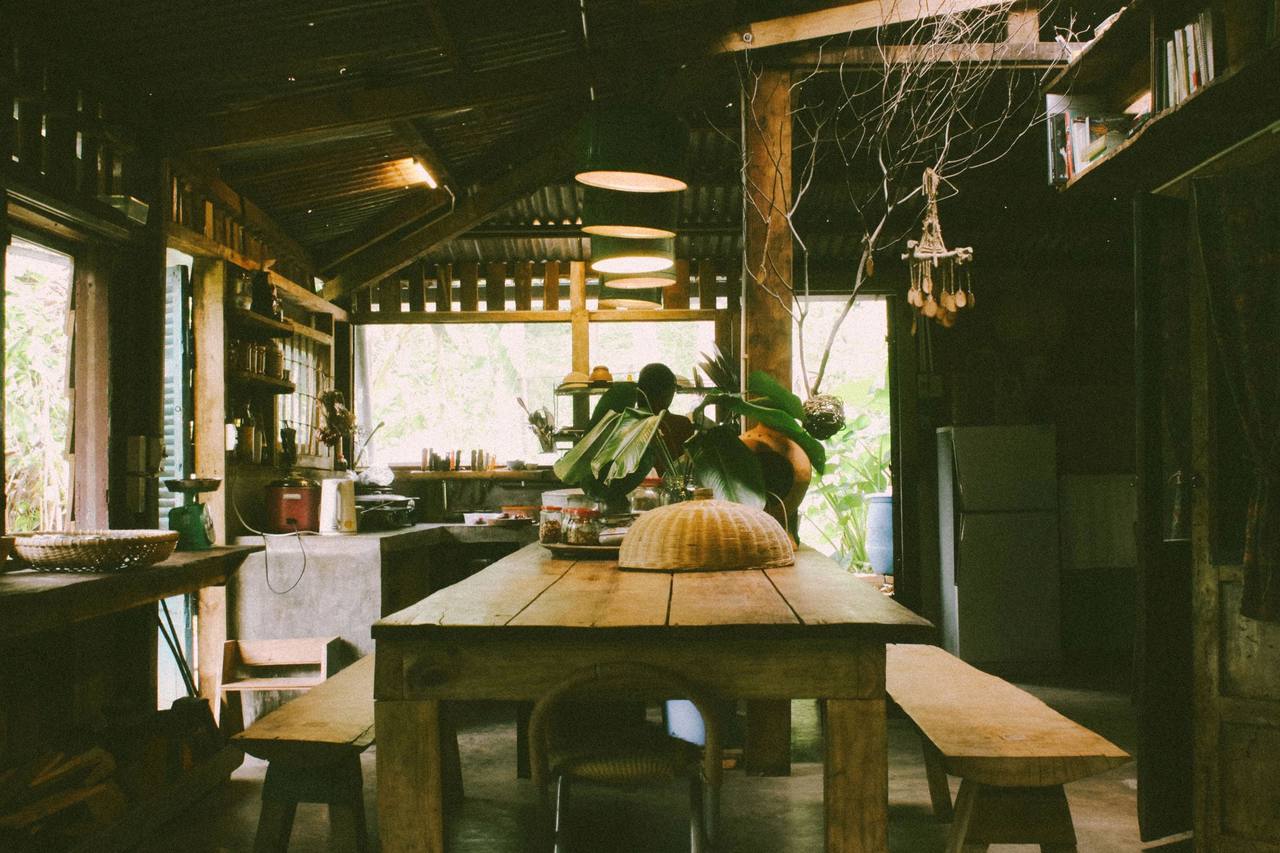 The Garden House kitchen in a timber house: a long wooden table with benches on both sides and a woven food cover, a cement counter with a rice cooker and a kettle, someone cooking at the back with their back turned, round pendant lamps under the roof, openings onto a green garden