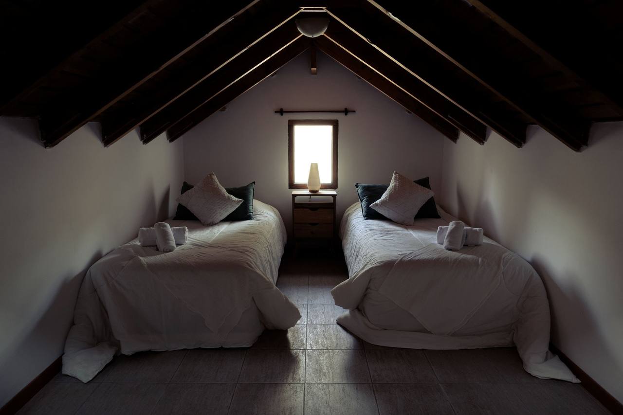 The Garden House second bedroom under the roof: two single beds with white duvets and rolled towels, a wooden bedside table with a lamp between them, a small wood-framed window, dark timber rafters over white walls