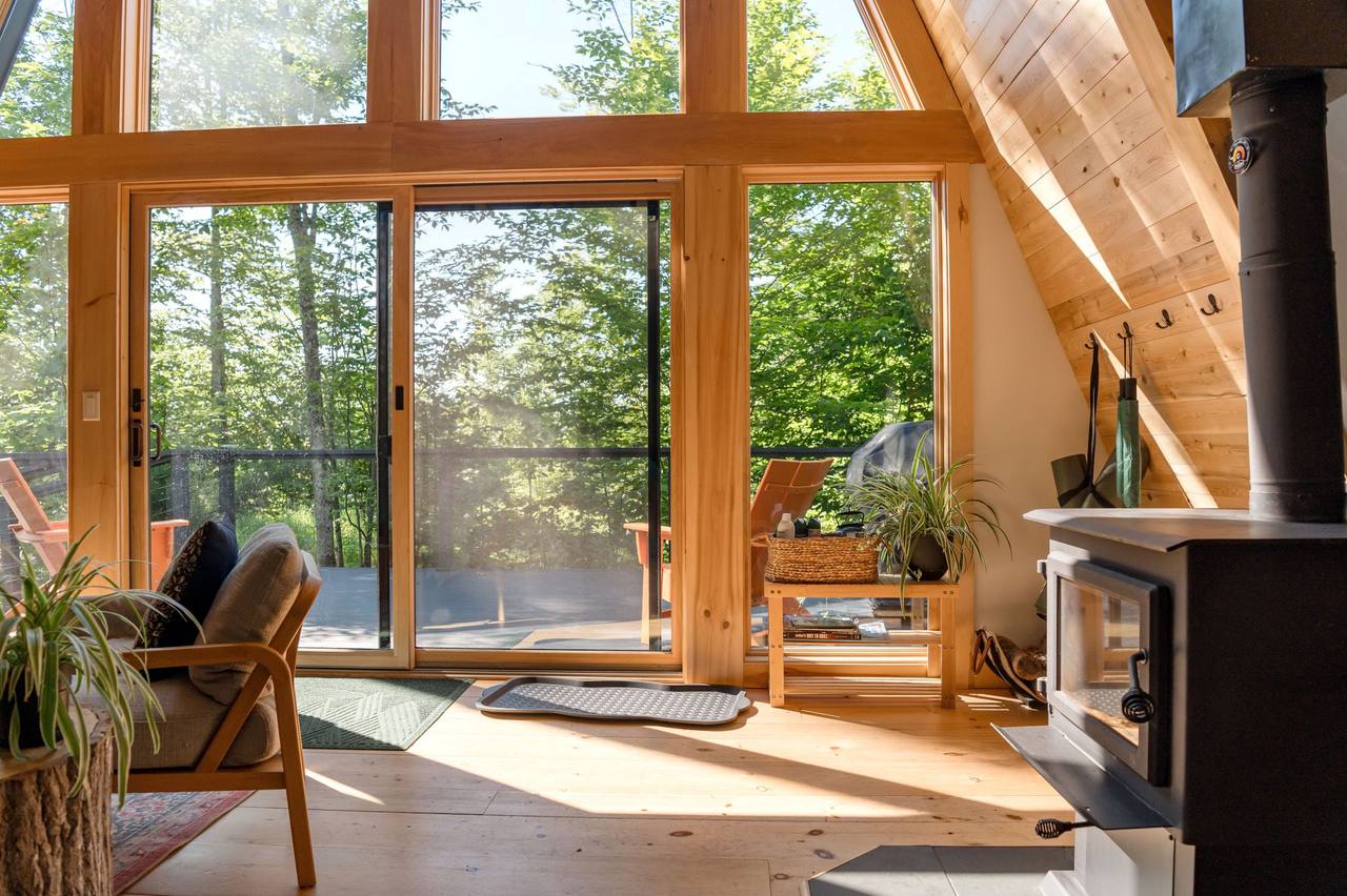 Garden House living room: a black wood stove on the right, a wooden armchair and plants on the left, sliding glass doors opening to the terrace and green woods