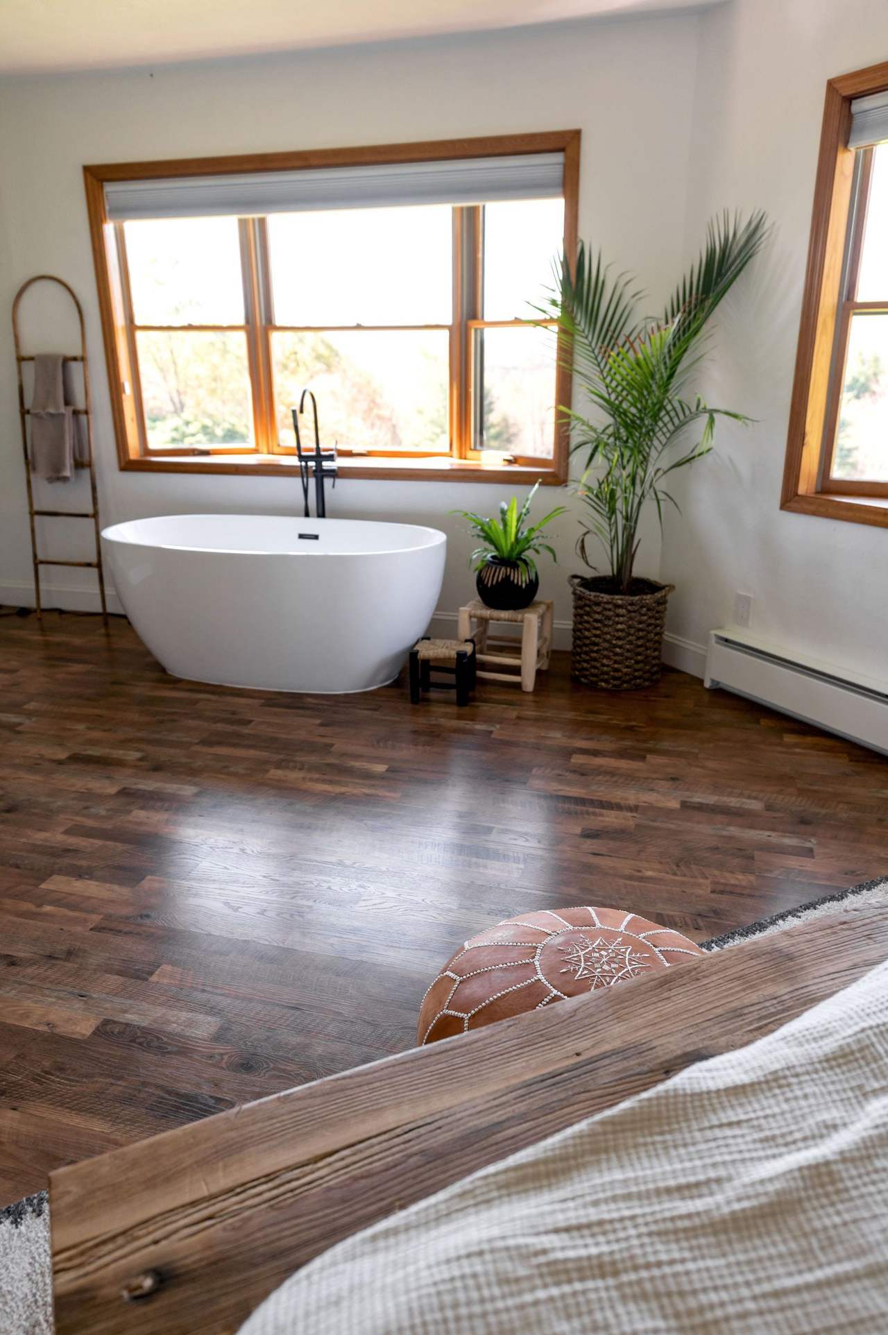 A white freestanding bath on the wooden floor right under a wood-framed window, a potted palm and a towel ladder beside it, the corner of the bed in the foreground