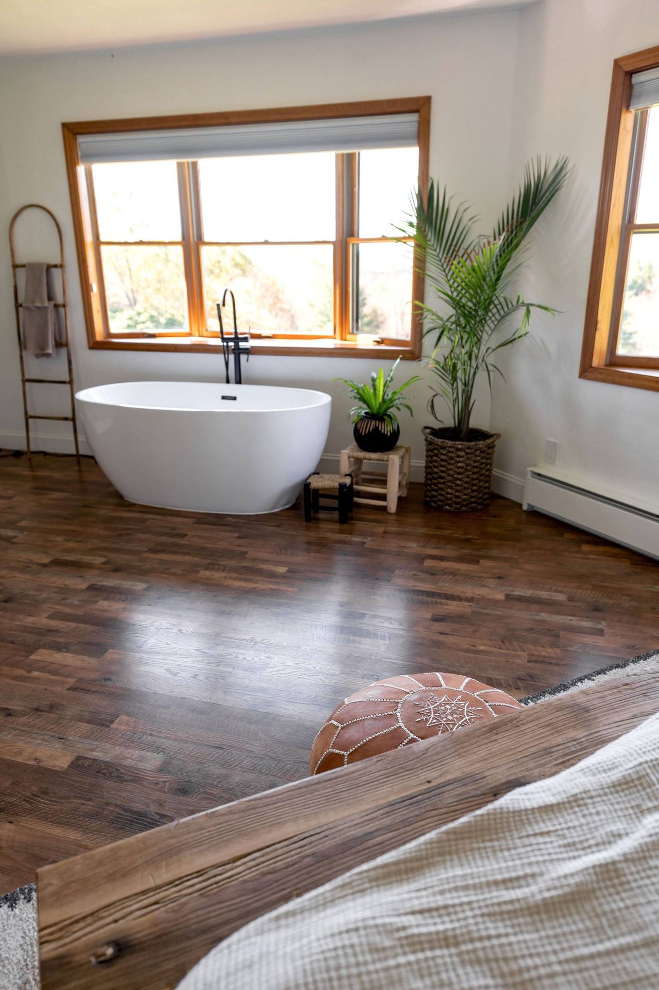 A white freestanding bath on the wooden floor right under a wood-framed window, a potted palm and a towel ladder beside it, the corner of the bed in the foreground