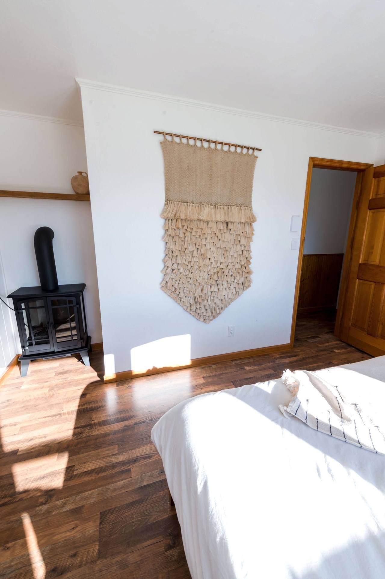 A black cast-iron wood stove in the corner of the Mist Suite, a macramé hanging on the white wall, sun slanting across the wooden floor and the edge of the bed