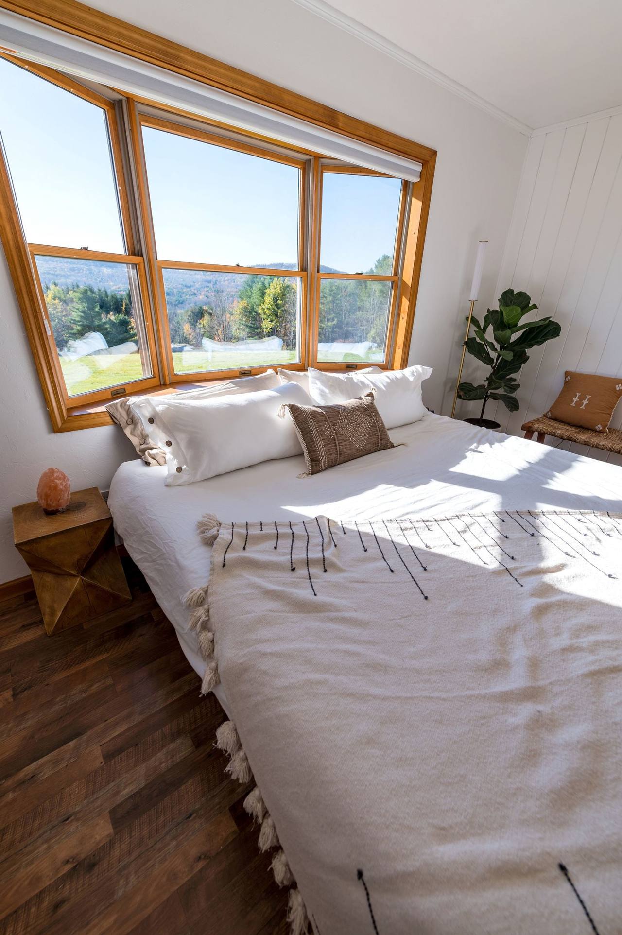 Mist Suite: a large bed in white linen under a row of wood-framed windows, early sun across the blanket, trees and distant hills outside