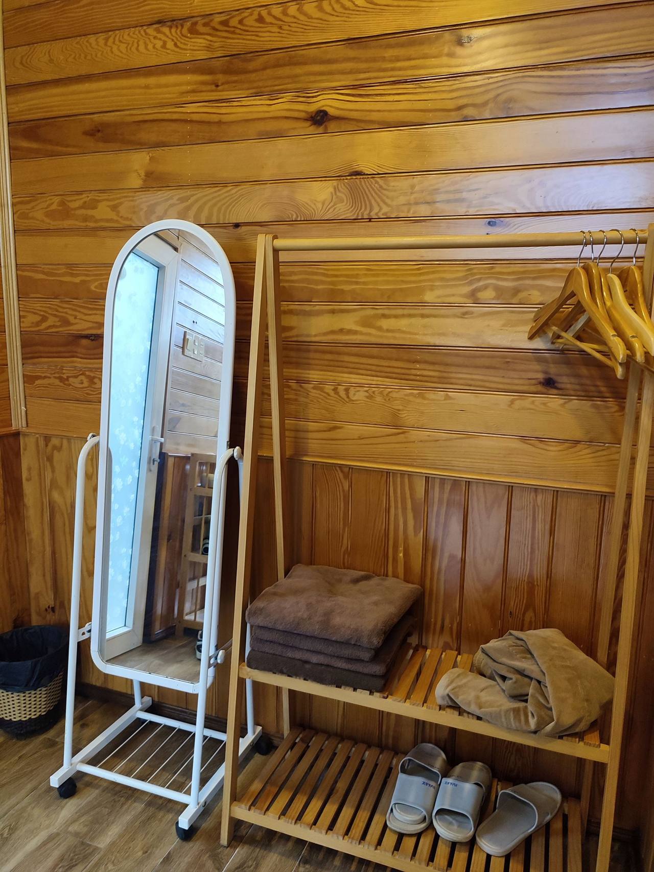 A corner of the Twin Pine room: a white standing mirror, a wooden rack with hangers, folded bath towels and two pairs of house slippers, a pine-board wall behind