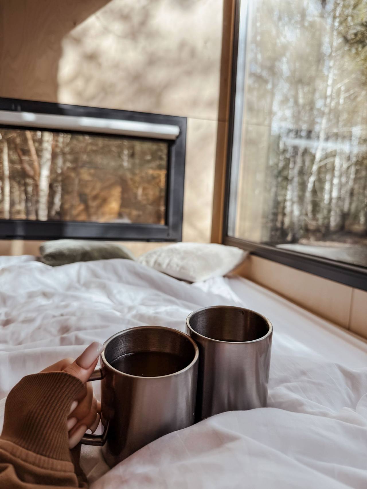 Little Pine: a hand in a knitted sleeve holds two metal mugs on the white sheet, the window behind looks out on the trees